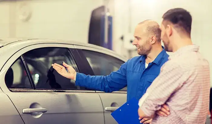 A technician explains the side window replacement process to a customer beside a silver car in a professional auto repair shop.